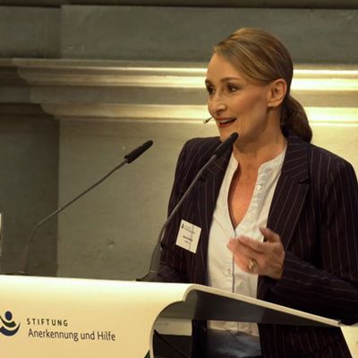 Moderatorin Sandra Olbrich in dunklem Blazer hinter einem Pult mit Mikrofonen bei einer Veranstaltung am 13.5.19 in Berlin