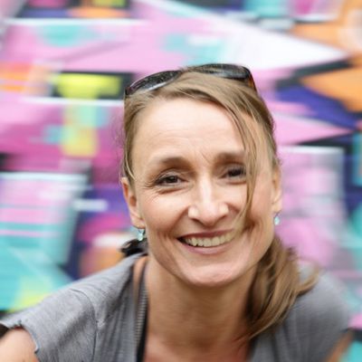 Portrait Sandra Olbrich, lachend und mit einer Haarsträhne im Gesicht, sie trägt ein graues T-Shirt, im Hintergrund eine bunte Graffiti-Wand