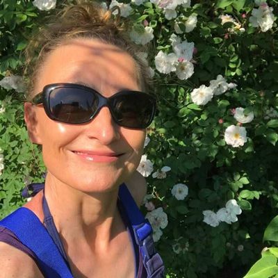 Sandra in blauer Gärtnerinnen-Latzhose und mit Sonnenbrille vor einer weißen Kletterrose in ihrem sonnigen Garten